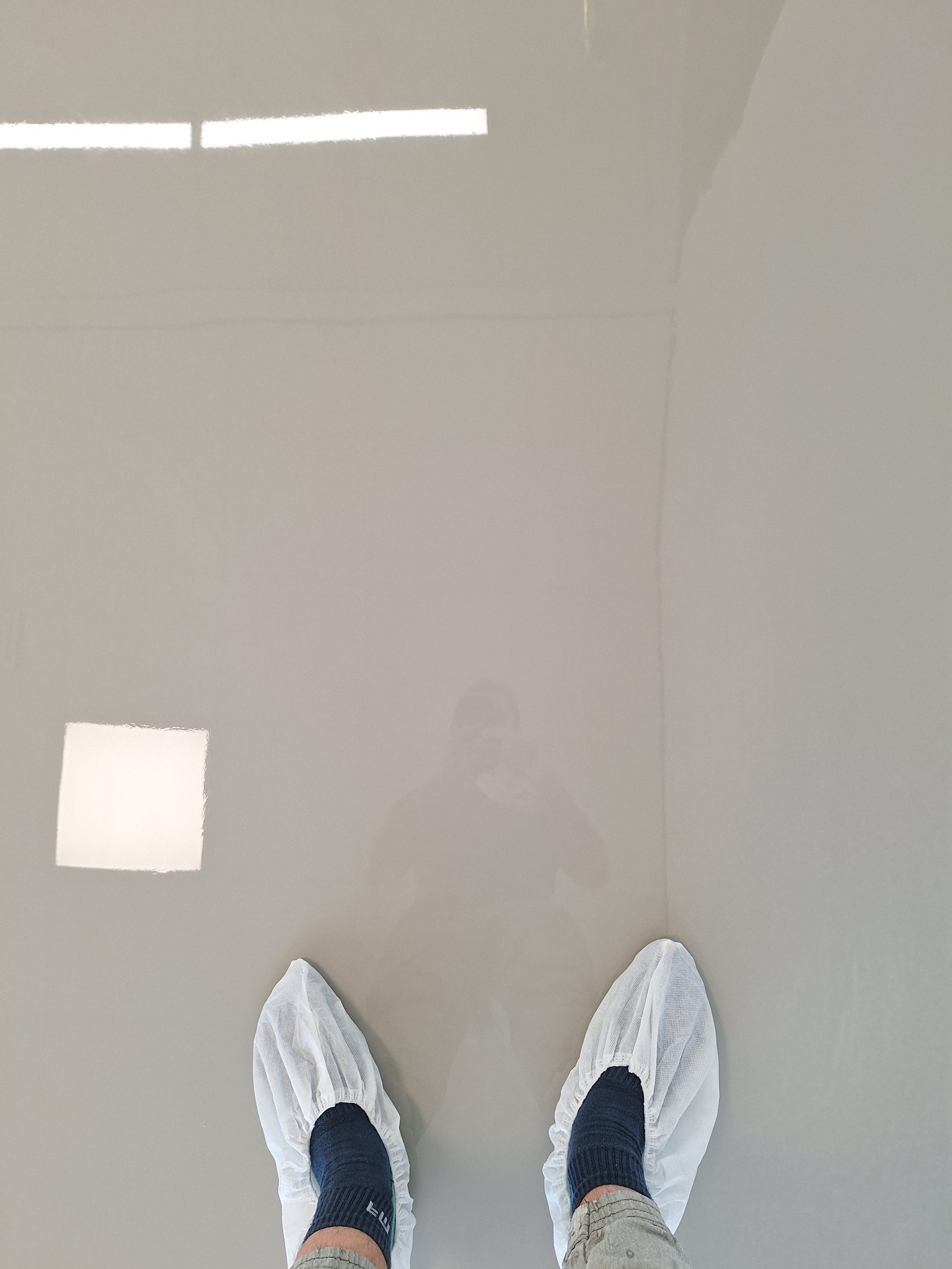 Świeżo wylana, jasnoszara posadzka z żywicy epoksydowej, widoczne odbicie światła i sylwetka osoby w ochronnych nakładkach na obuwiu.