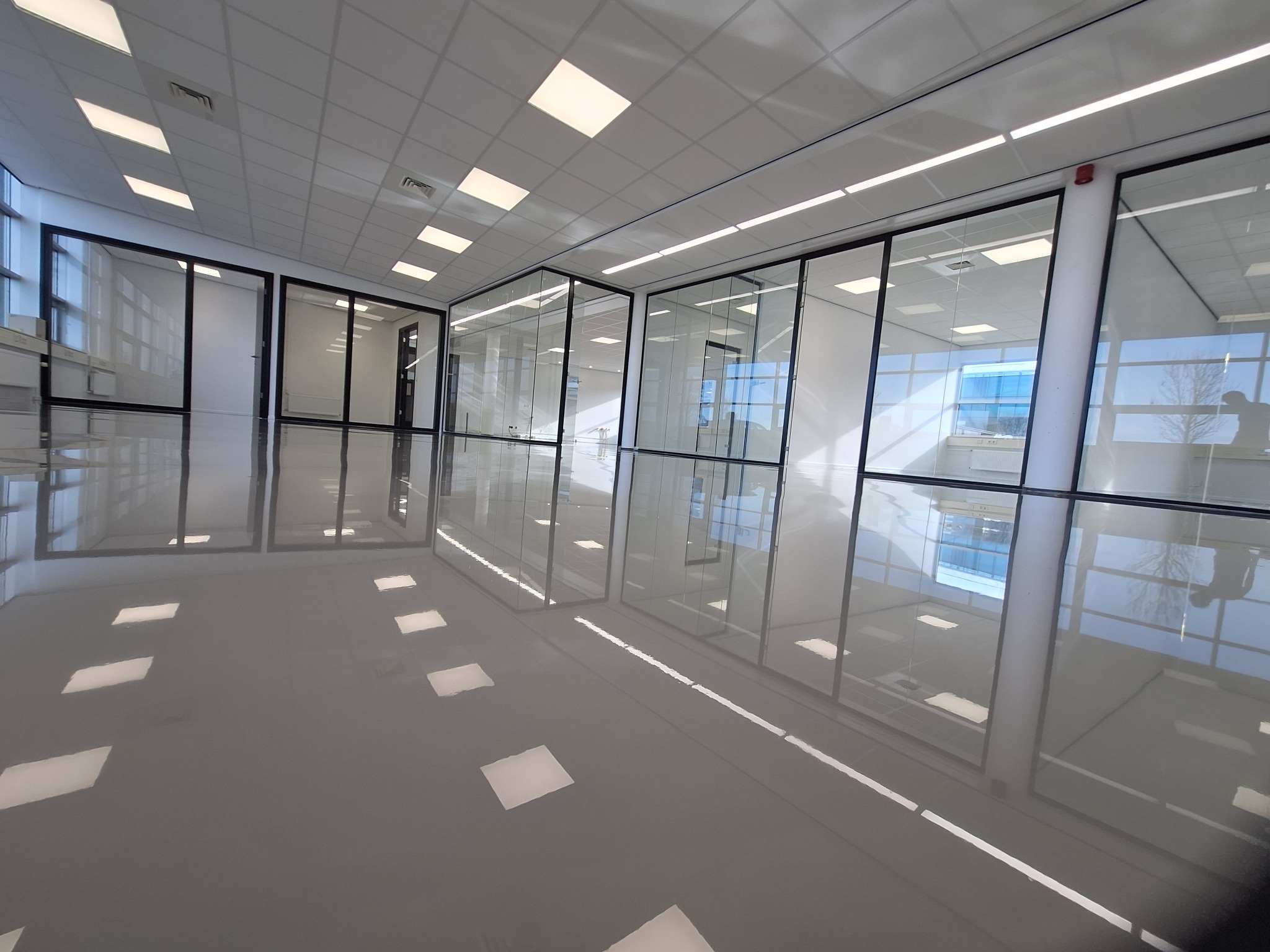 Błyszcząca, szara posadzka żywiczna w przestronnym biurze z przeszklonymi ścianami działowymi, odbijająca światło sufitowych lamp.
