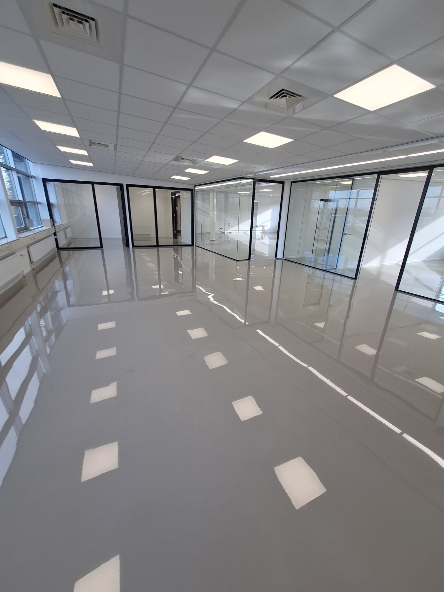 Jasnoszara, błyszcząca posadzka w biurze z przeszklonymi ścianami działowymi, widoczne odbicia oświetlenia sufitowego.