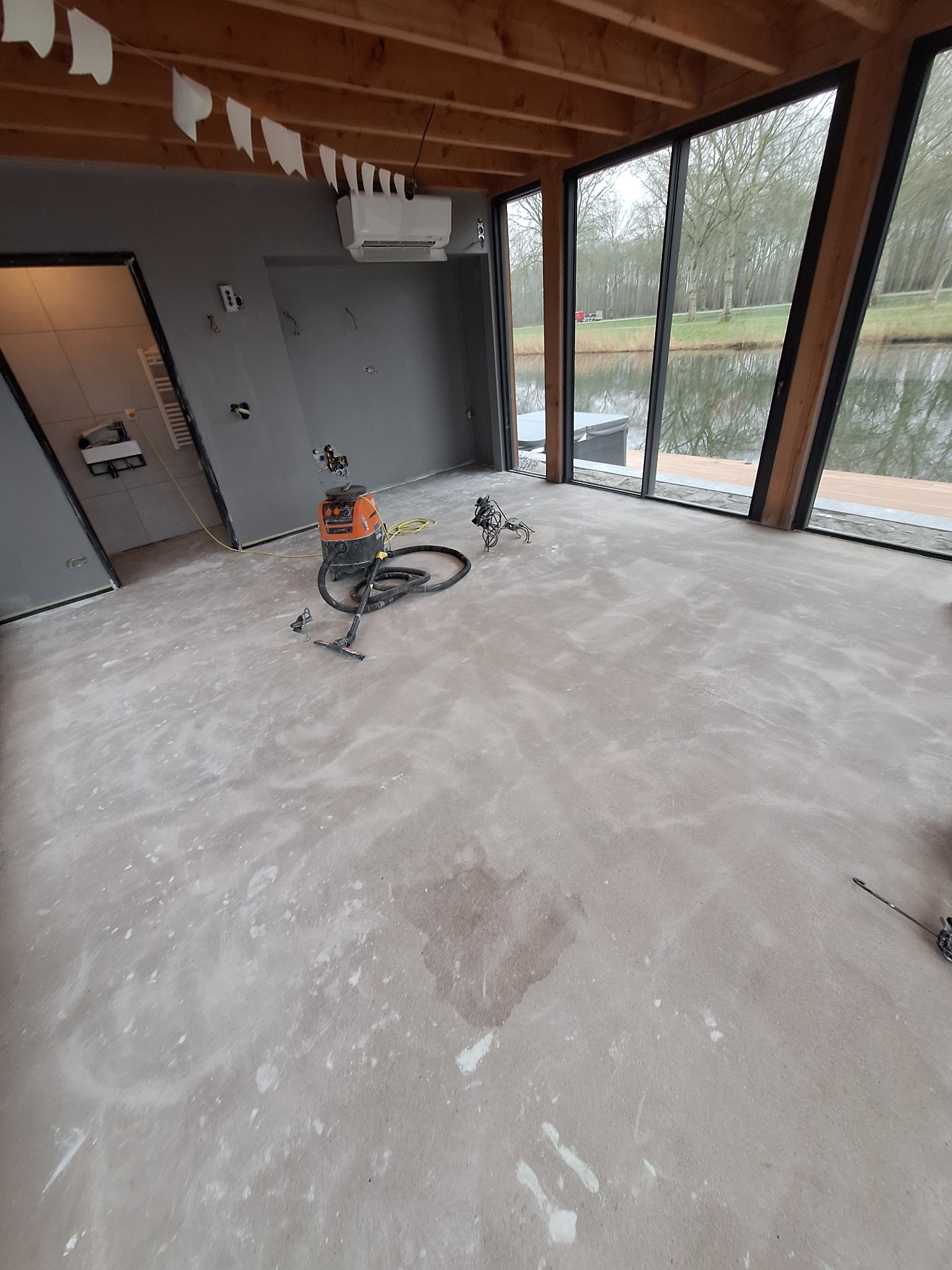 Wnętrze pomieszczenia z dużymi oknami i widokiem na kanał, podłoga w trakcie prac wykończeniowych, widoczny odkurzacz przemysłowy i ślady po szpachlowaniu.