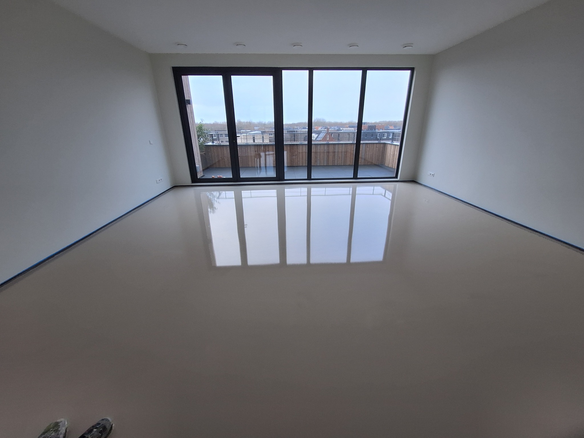 Jasne, przestronne wnętrze z beżową, błyszczącą posadzką żywiczną, odbijającą światło z dużego okna balkonowego z widokiem na zabudowę miejską. W rogu widoczne narzędzia malarskie.