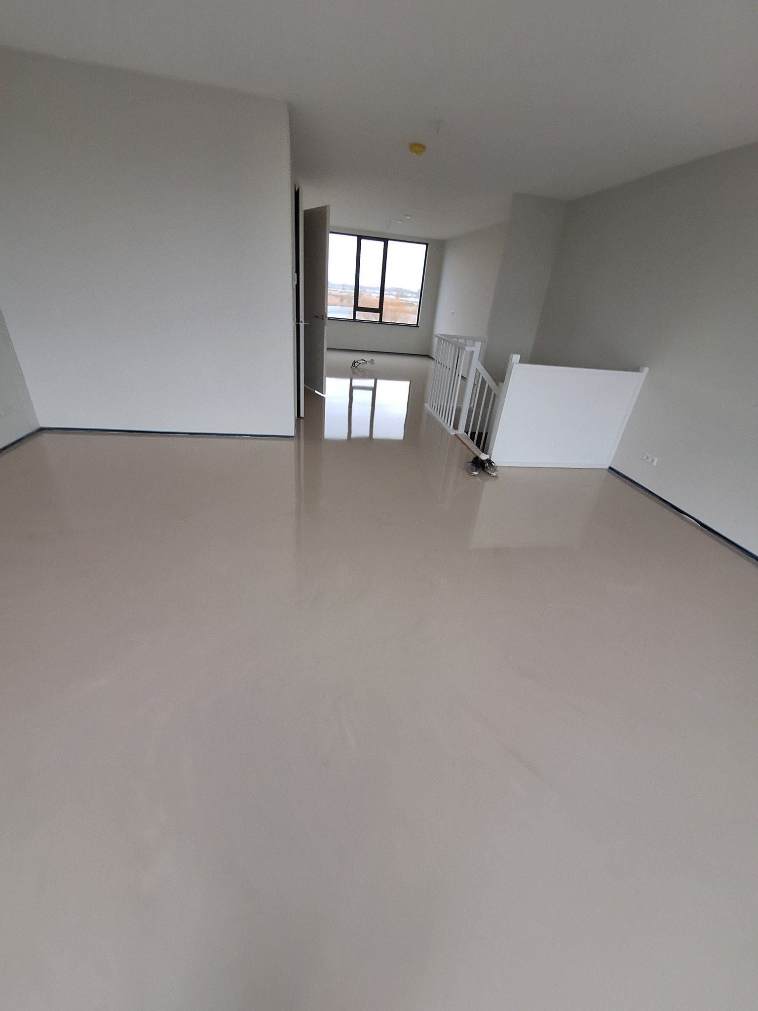 Wnętrze wykończonego, pustego pokoju z jasną, błyszczącą posadzką i białymi ścianami, widoczne schody z białą balustradą, przez okno widać budynki.