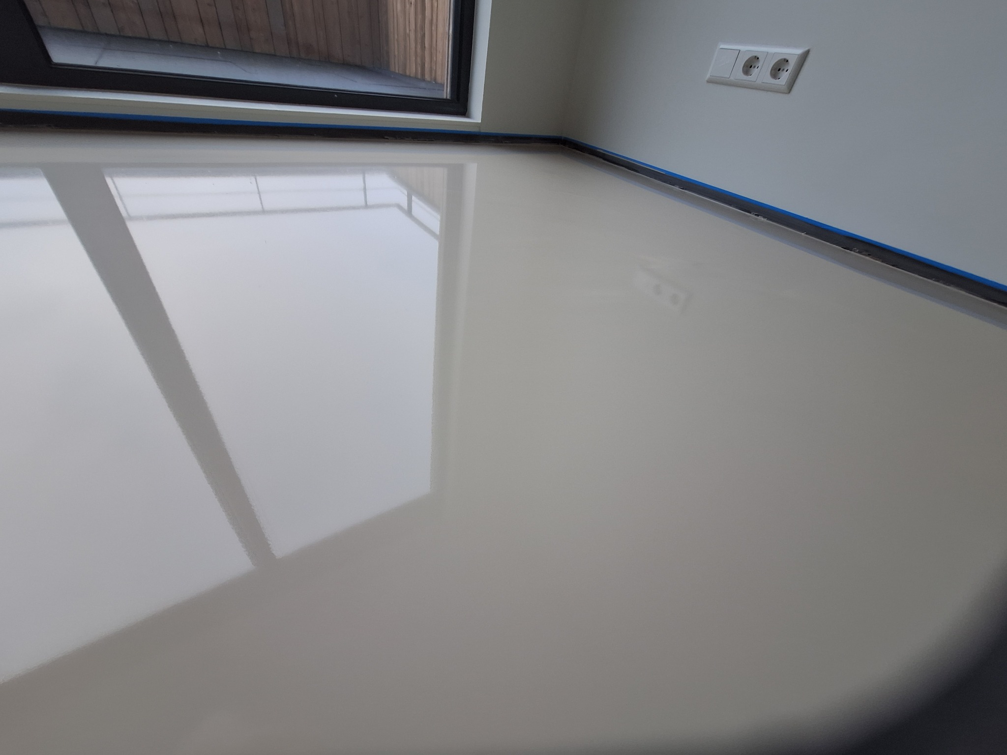 Beżowa, błyszcząca posadzka w nowym pomieszczeniu z oknem i gniazdkiem elektrycznym na ścianie, zabezpieczona taśmą malarską przy listwach przypodłogowych i oknie.