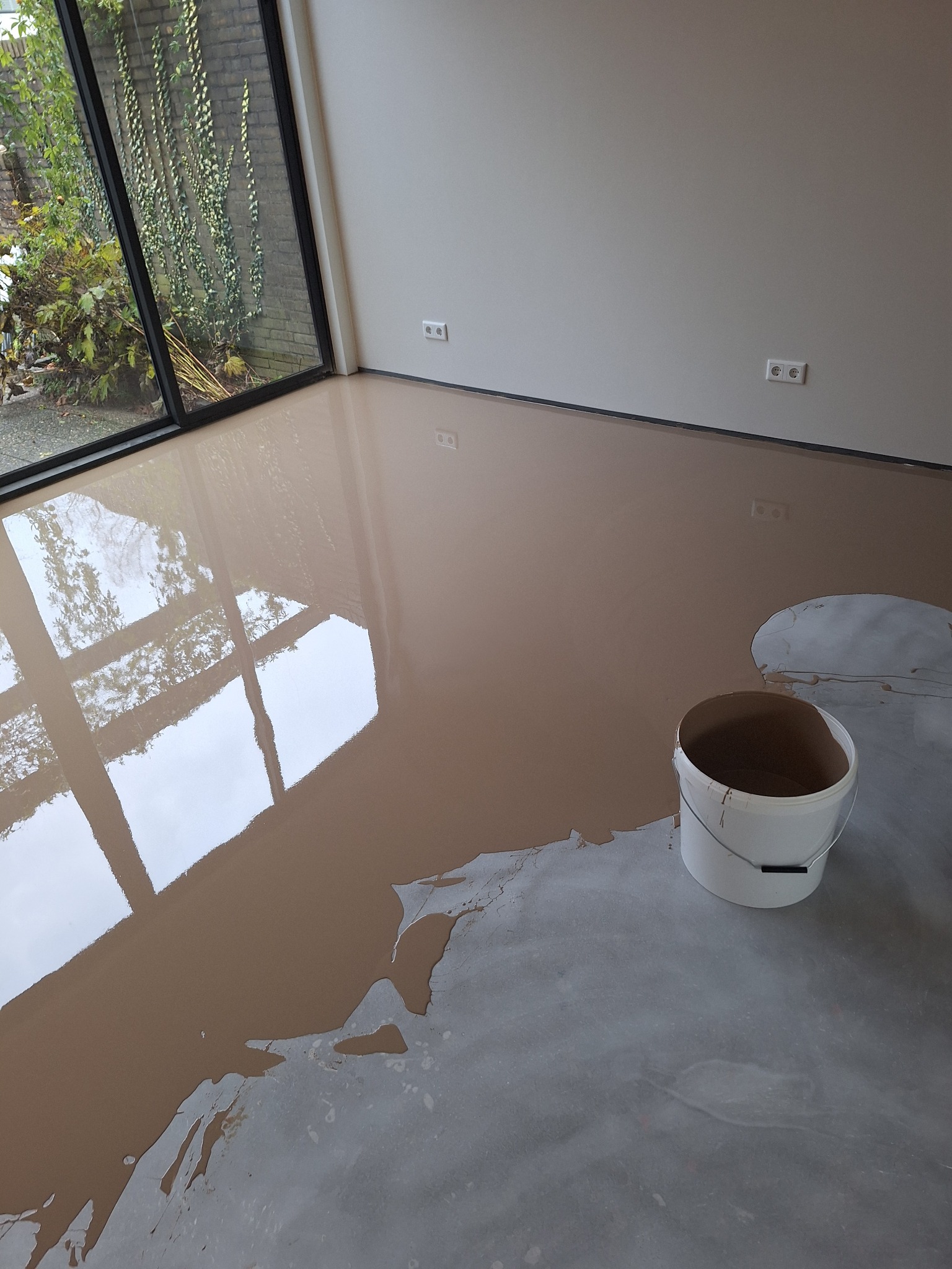 Świeżo wylana, beżowa posadzka samopoziomująca w jasnym pomieszczeniu z dużym oknem, wiadro z resztkami masy obok