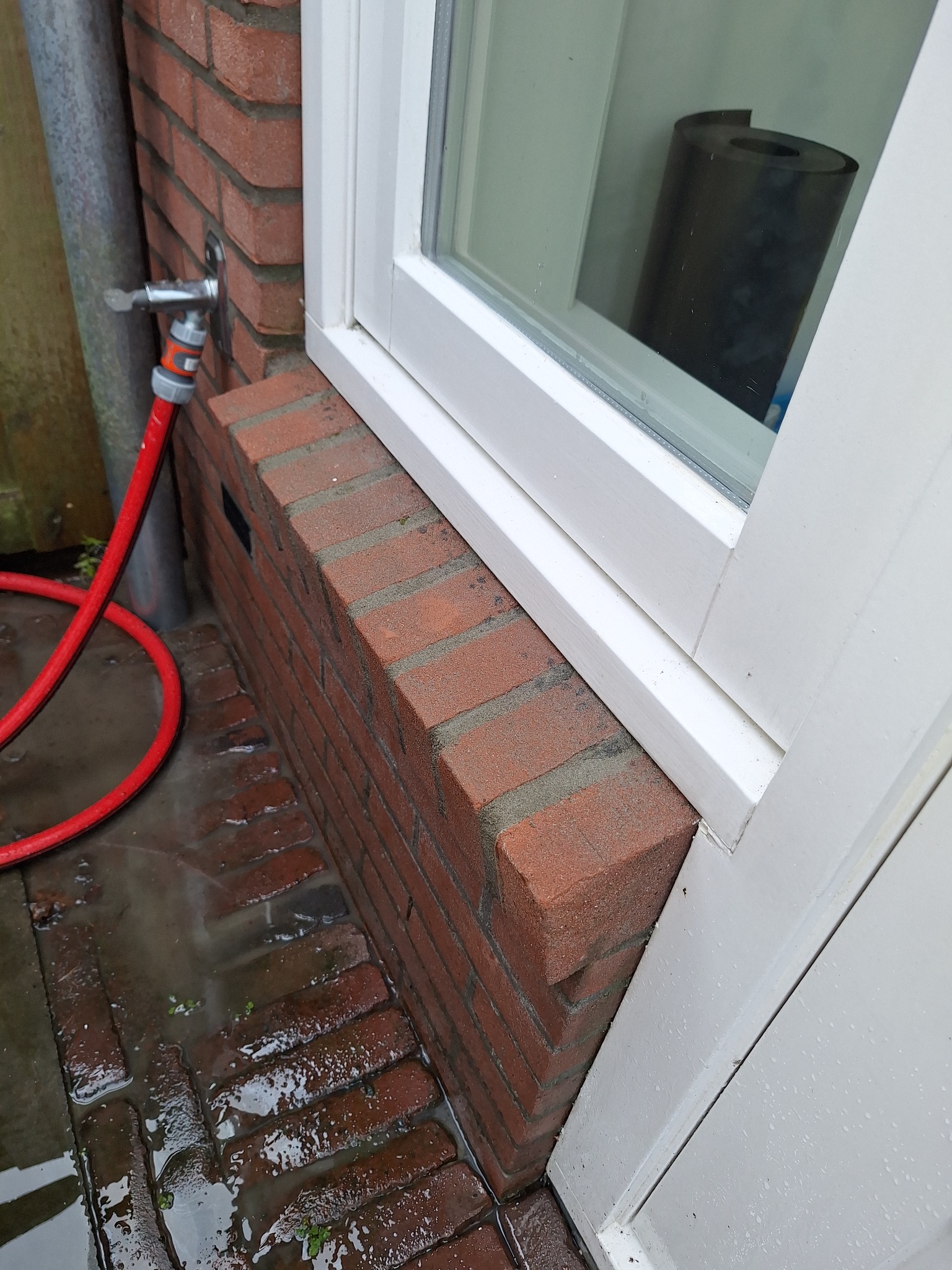 Murowany parapet zewnętrzny z czerwonej cegły pod białym oknem, obok ściany z kranem i czerwoną rurą ogrodową, na mokrym brukowanym podłożu.