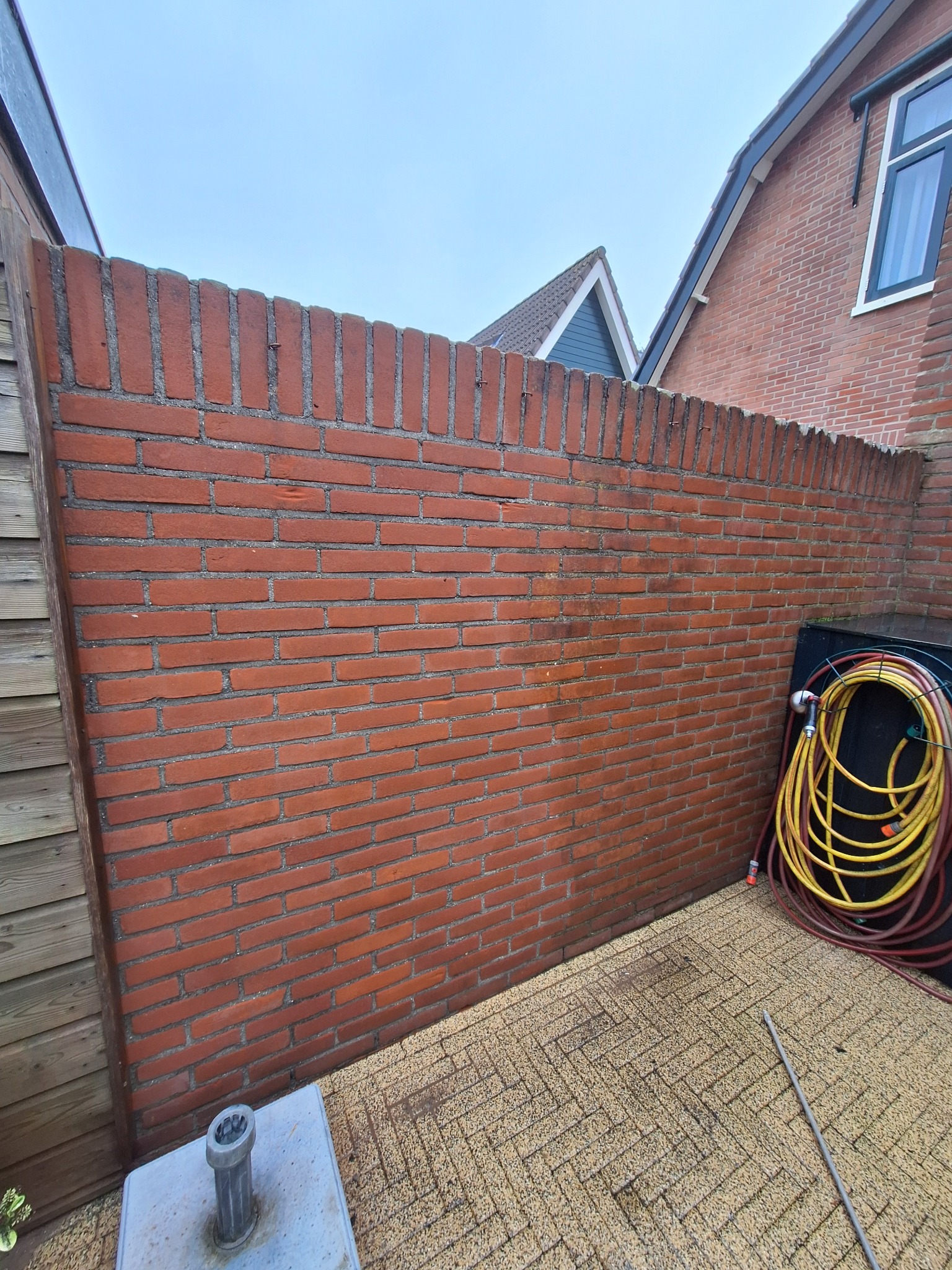 Ceglany mur ogrodzeniowy z wykończeniem z cegieł pionowych, widoczny fragment domu sąsiada i zwinięty wąż ogrodowy.