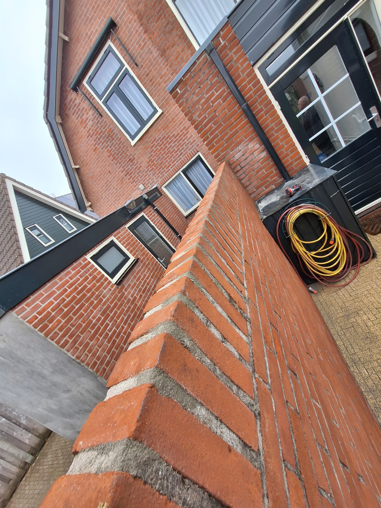 Fragment muru z czerwonej cegły w Alphen aan den Rijn, widoczny z bliska pod nietypowym kątem, z fragmentami budynku w tle i zwiniętym wężem ogrodowym obok drzwi.