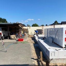 Usługi remontowo budowlane Jabczyński szczepan - Pracownik w niebieskiej koszulce szlifuje bloczki z betonu komórkowego Ytong na fundamencie w trakcie budowy domu, w tle zapakowane palety z bloczkami, widoczne narzędzia i elementy placu budowy.