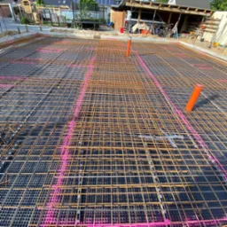 Przygotowanie zbrojenia płyty fundamentowej z rurami instalacyjnymi, widoczne siatki stalowe i folia kubełkowa z zaznaczonym r&oacute;żowym sprayem układem zbrojenia.