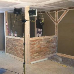 Usługi remontowo budowlane Jabczyński szczepan - Wnętrze w trakcie wykończenia: ściana z cegły klinkierowej do połowy wysokości, oświetlenie budowlane na statywie, pracownik tynkujący ścianę w tle, sufit z płyt karton-gips.