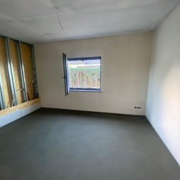 Usługi remontowo budowlane Jabczyński szczepan - Surowe wnętrze w Olszynie: Świeżo wylana, szara podłoga, ściany przygotowane do malowania, widoczne puszki elektryczne i częściowo odsłonięta konstrukcja ściany z metalowymi profilami.