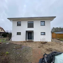 Nowoczesny, dwukondygnacyjny dom z płaskim dachem i białymi ścianami, z czarnymi ramami okiennymi i drzwiami wejściowymi, w trakcie prac wykończeniowych ogrodu, widoczne krokusy i przygotowane rabaty.