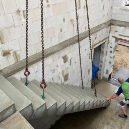 Usługi remontowo budowlane Jabczyński szczepan - Montaż prefabrykowanych betonowych schodów na budowie domu z bloczków, podwieszonych na stalowych łańcuchach z hakami, w tle ściany z betonu komórkowego.