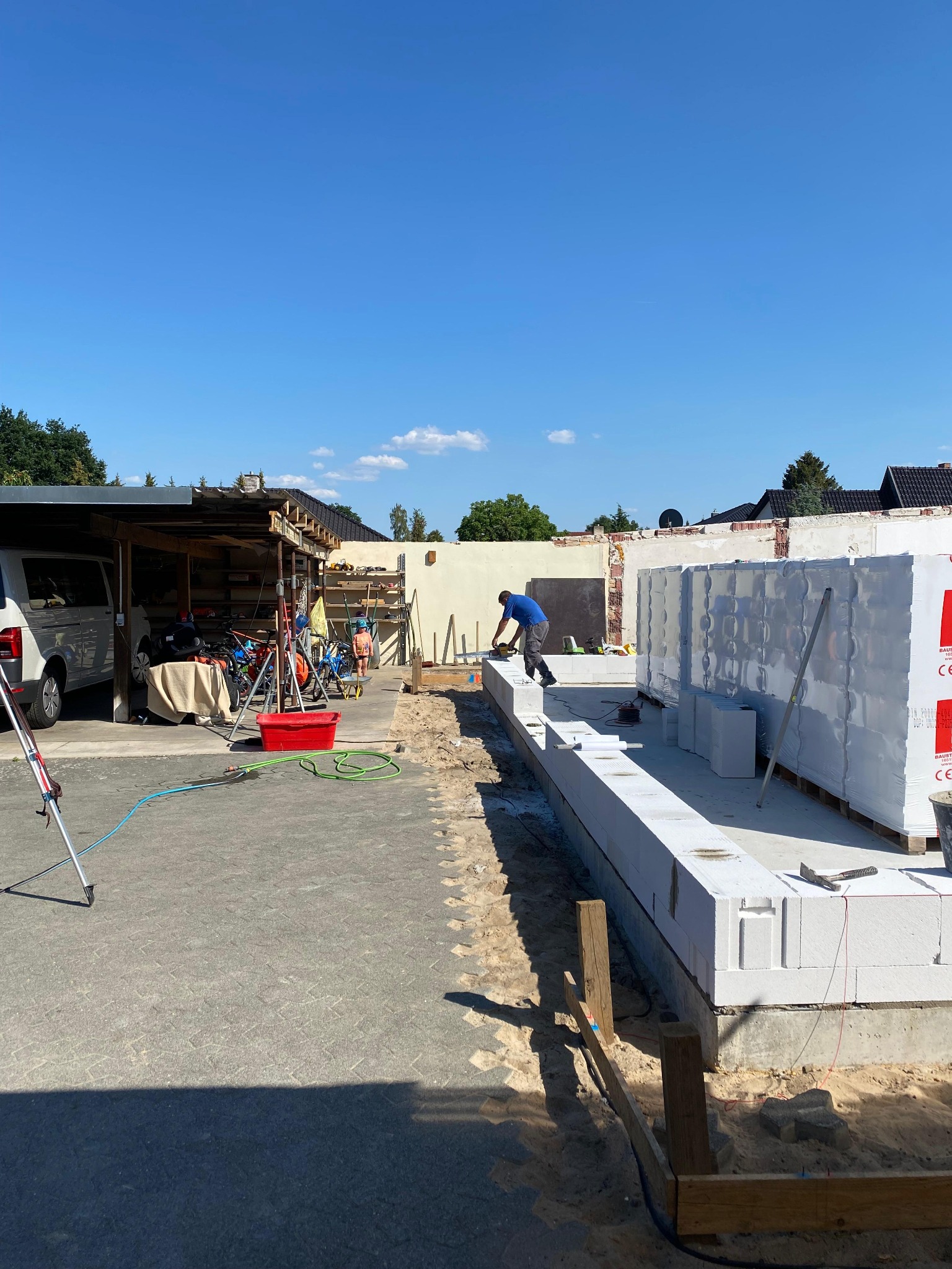 Pracownik w niebieskiej koszulce szlifuje bloczki z betonu komórkowego Ytong na fundamencie w trakcie budowy domu, w tle zapakowane palety z bloczkami, widoczne narzędzia i elementy placu budowy.