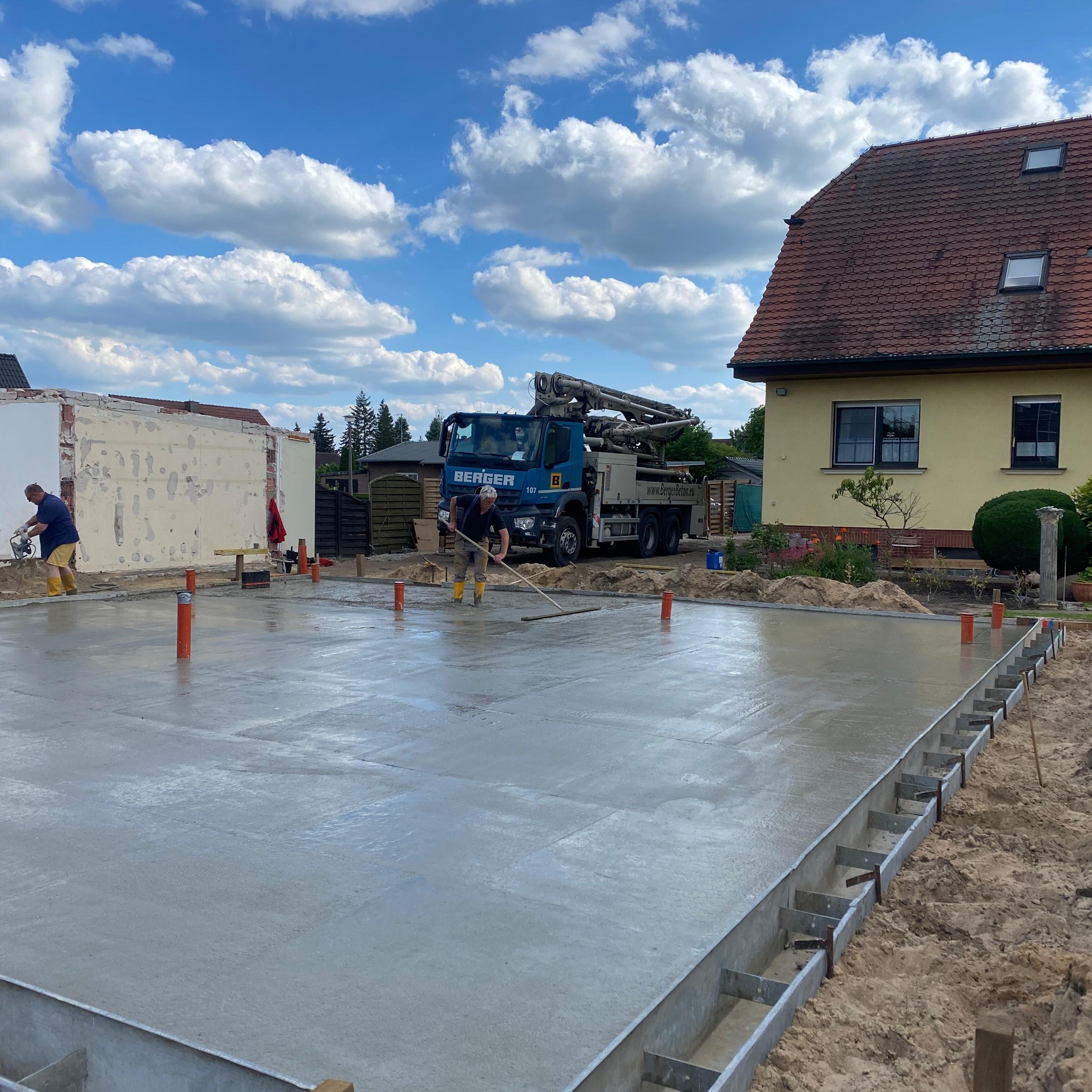 Wylewanie fundamentów pod budynek, pracownik wygładza powierzchnię betonu przy użyciu specjalnej listwy, w tle betonomieszarka firmy Berger i dom z czerwonym dachem.