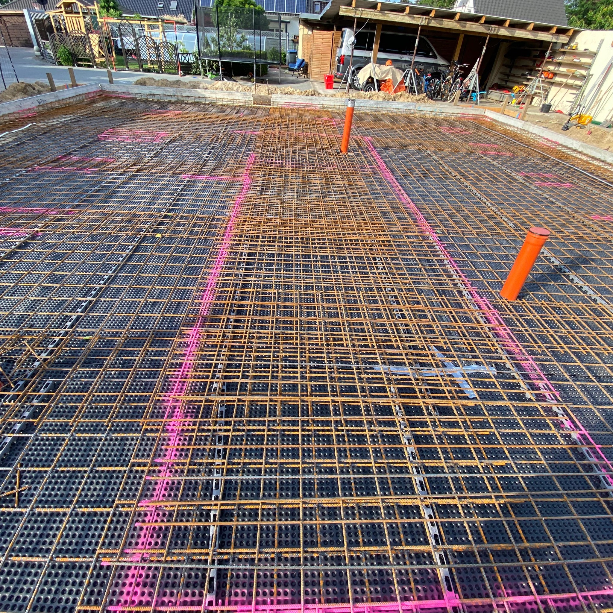 Przygotowanie zbrojenia płyty fundamentowej z rurami instalacyjnymi, widoczne siatki stalowe i folia kubełkowa z zaznaczonym różowym sprayem układem zbrojenia.
