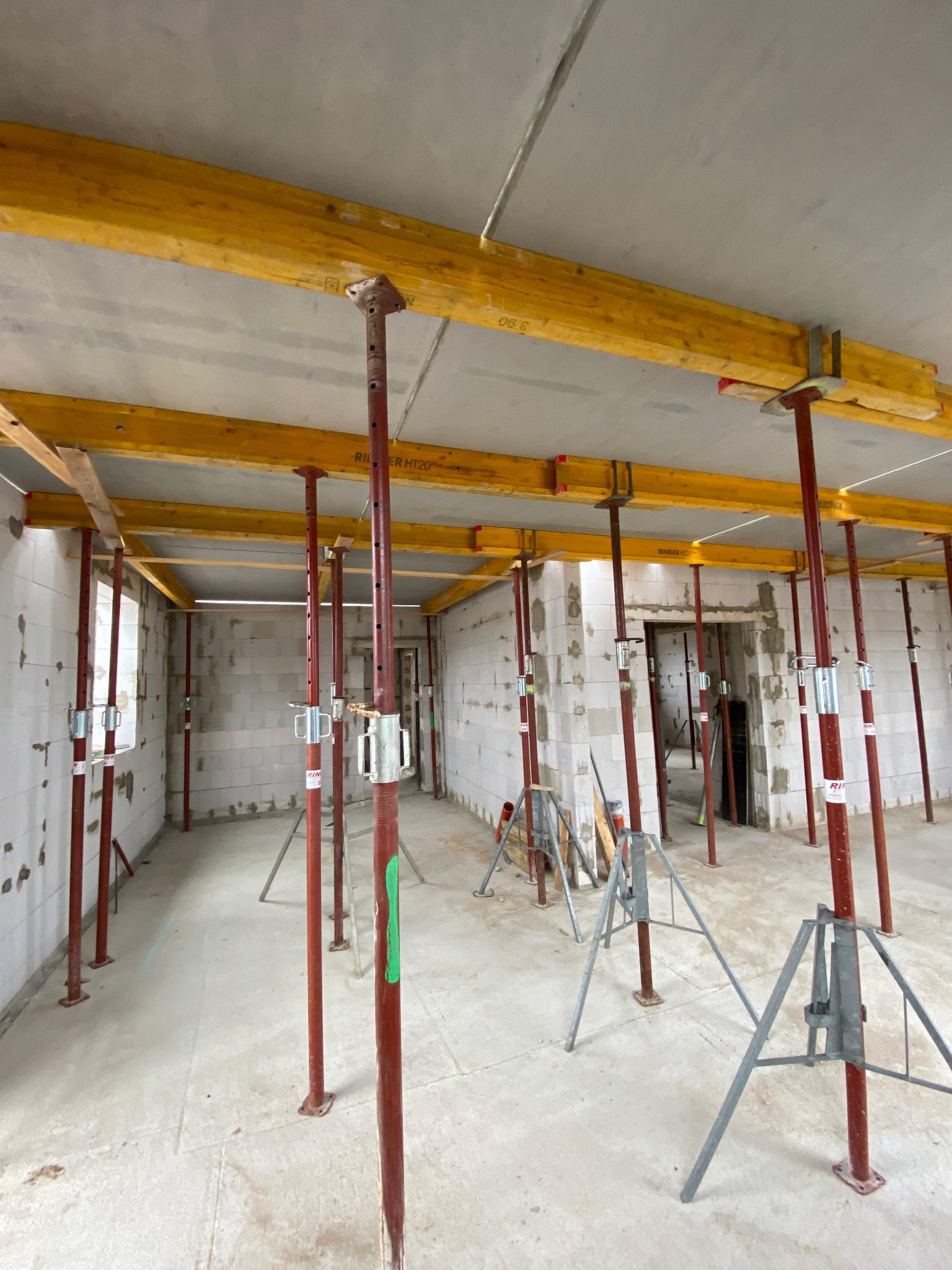 Wnętrze domu w trakcie budowy z białych bloczków, z widocznymi stemplami podtrzymującymi strop i żółtymi belkami, podłoga betonowa, widoczne okno i otwór drzwiowy.