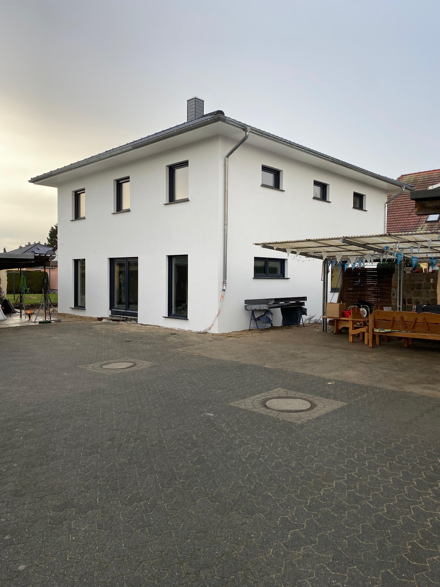 Nowoczesny, dwukondygnacyjny dom murowany z białymi ścianami zewnętrznymi i ciemnymi ramami okiennymi, widoczny z poziomu brukowanego podjazdu, z elementami wykończeniowymi wciąż w trakcie realizacji.