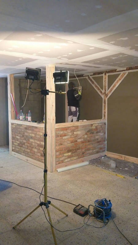 Wnętrze w trakcie wykończenia: ściana z cegły klinkierowej do połowy wysokości, oświetlenie budowlane na statywie, pracownik tynkujący ścianę w tle, sufit z płyt karton-gips.