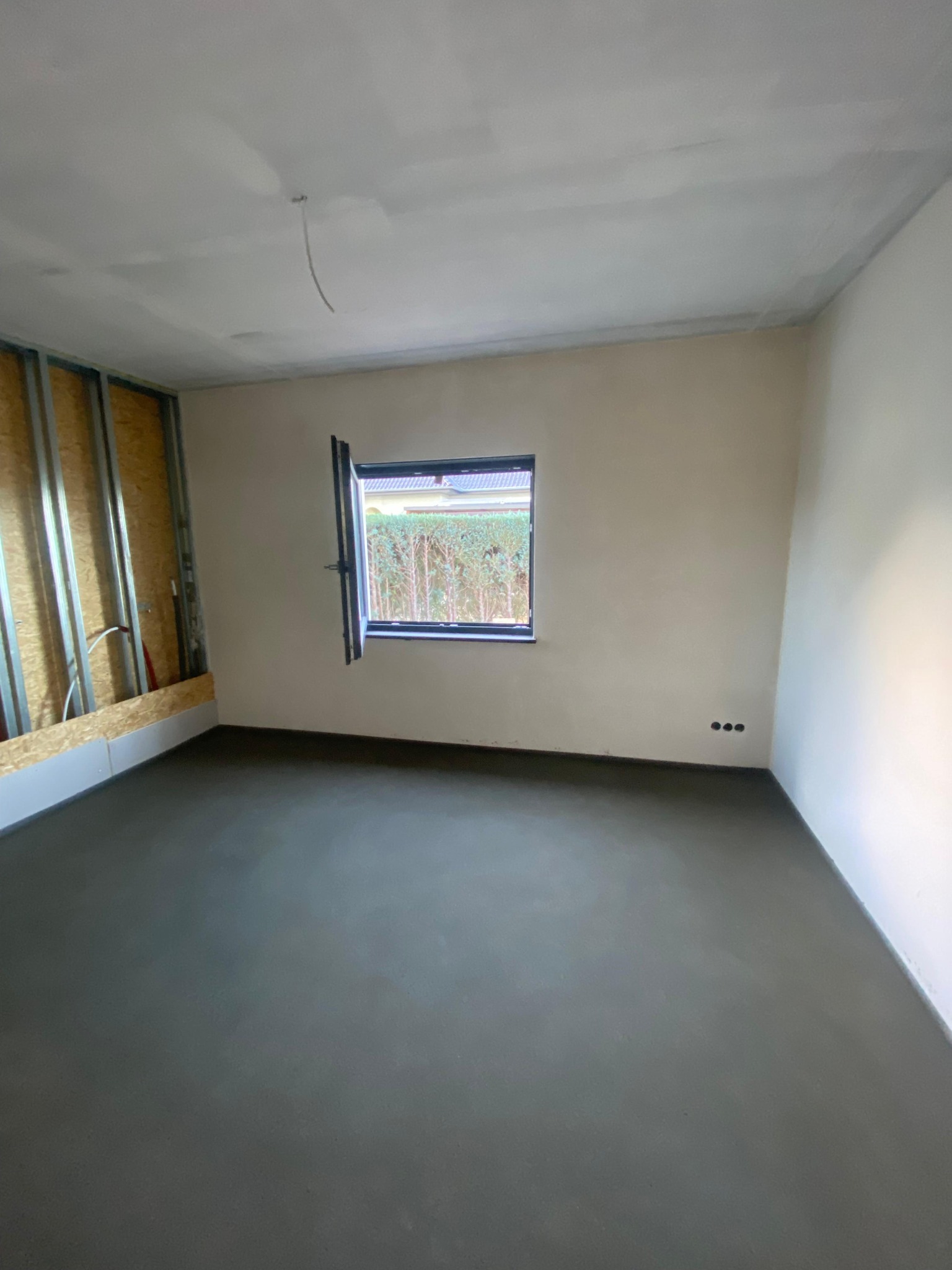 Surowe wnętrze w Olszynie: Świeżo wylana, szara podłoga, ściany przygotowane do malowania, widoczne puszki elektryczne i częściowo odsłonięta konstrukcja ściany z metalowymi profilami.