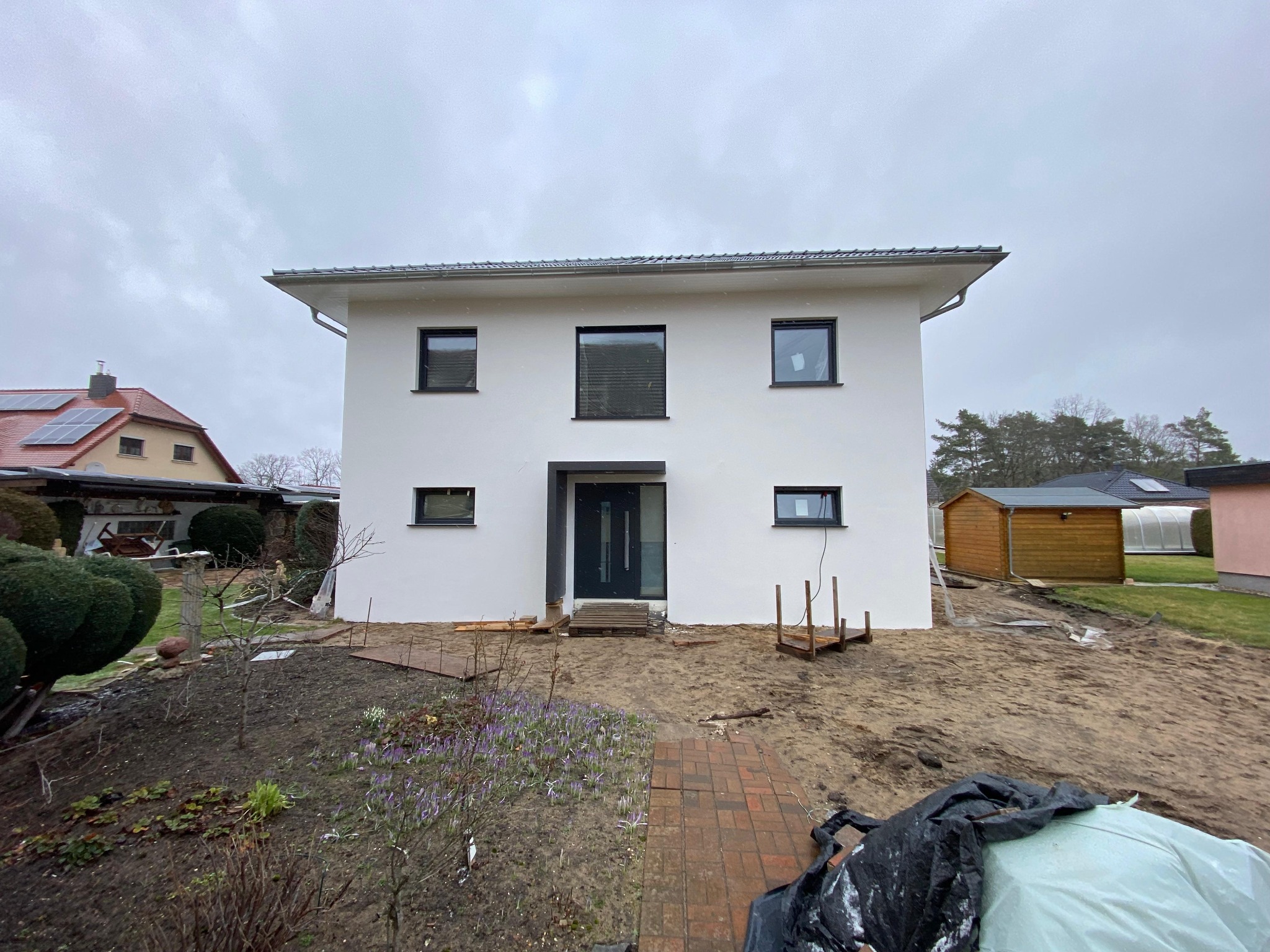 Nowoczesny, dwukondygnacyjny dom z płaskim dachem i białymi ścianami, z czarnymi ramami okiennymi i drzwiami wejściowymi, w trakcie prac wykończeniowych ogrodu, widoczne krokusy i przygotowane rabaty.