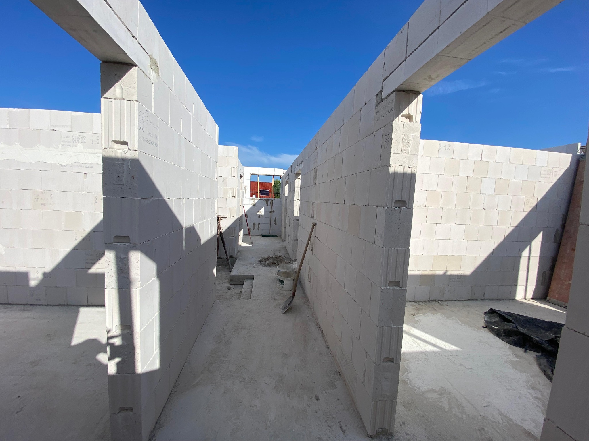 Wnętrze surowego domu z białych bloczków, widoczne ściany działowe i otwory okienne, na podłodze leżą narzędzia budowlane, w tle błękitne niebo.