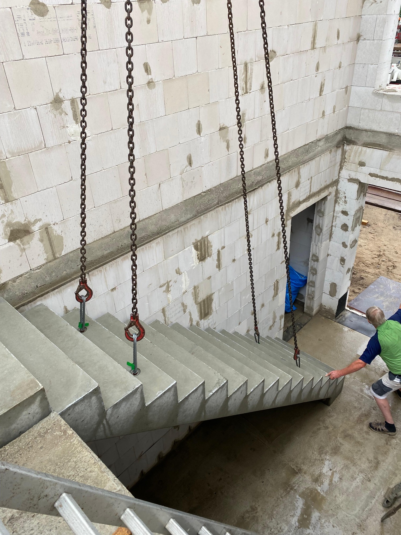 Montaż prefabrykowanych betonowych schodów na budowie domu z bloczków, podwieszonych na stalowych łańcuchach z hakami, w tle ściany z betonu komórkowego.