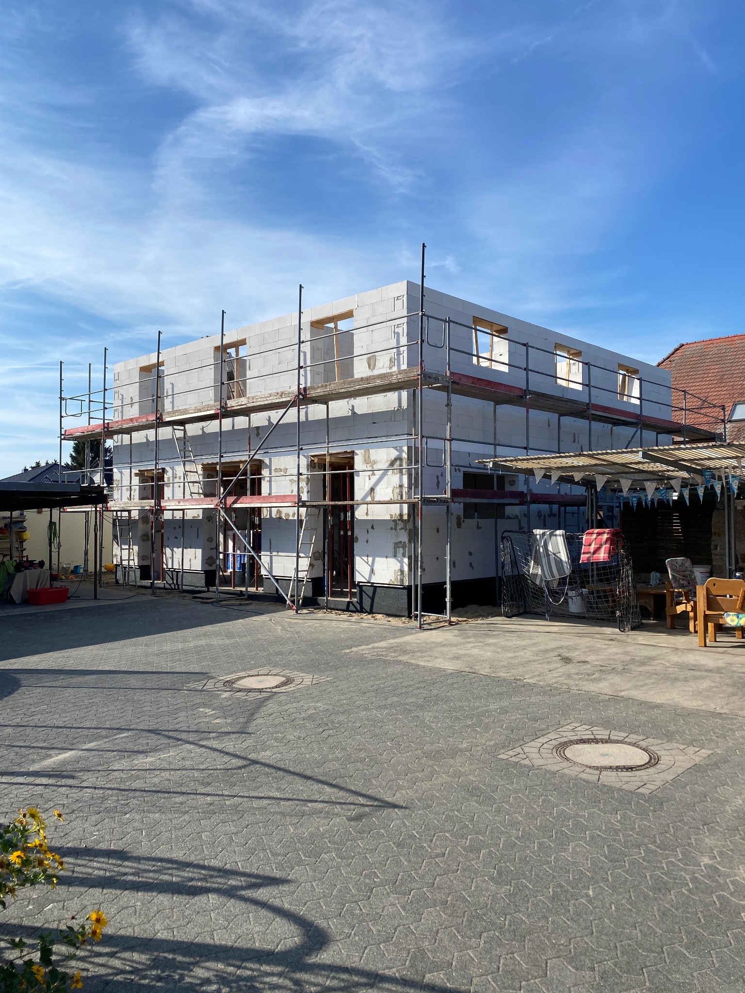 Dwukondygnacyjny budynek w trakcie wznoszenia z białych bloczków, otoczony rusztowaniami, widok z zewnątrz na tle błękitnego nieba. Na pierwszym planie kostka brukowa i żółte kwiaty.