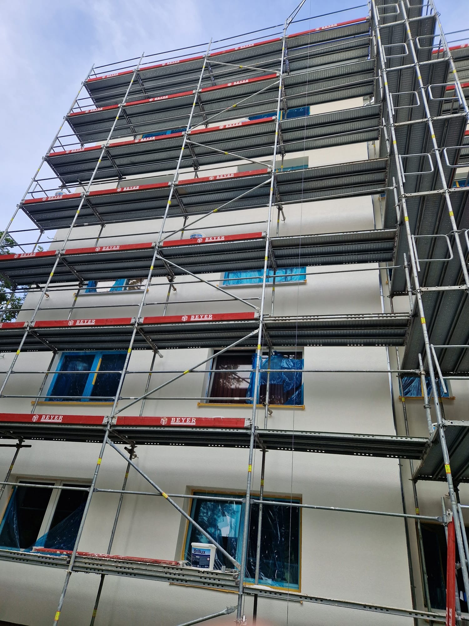 Wielopiętrowe rusztowanie z czerwonymi akcentami obejmuje białą elewację budynku mieszkalnego z oknami zabezpieczonymi folią, widoczny pojemnik z farbą na jednym z poziomów rusztowania.