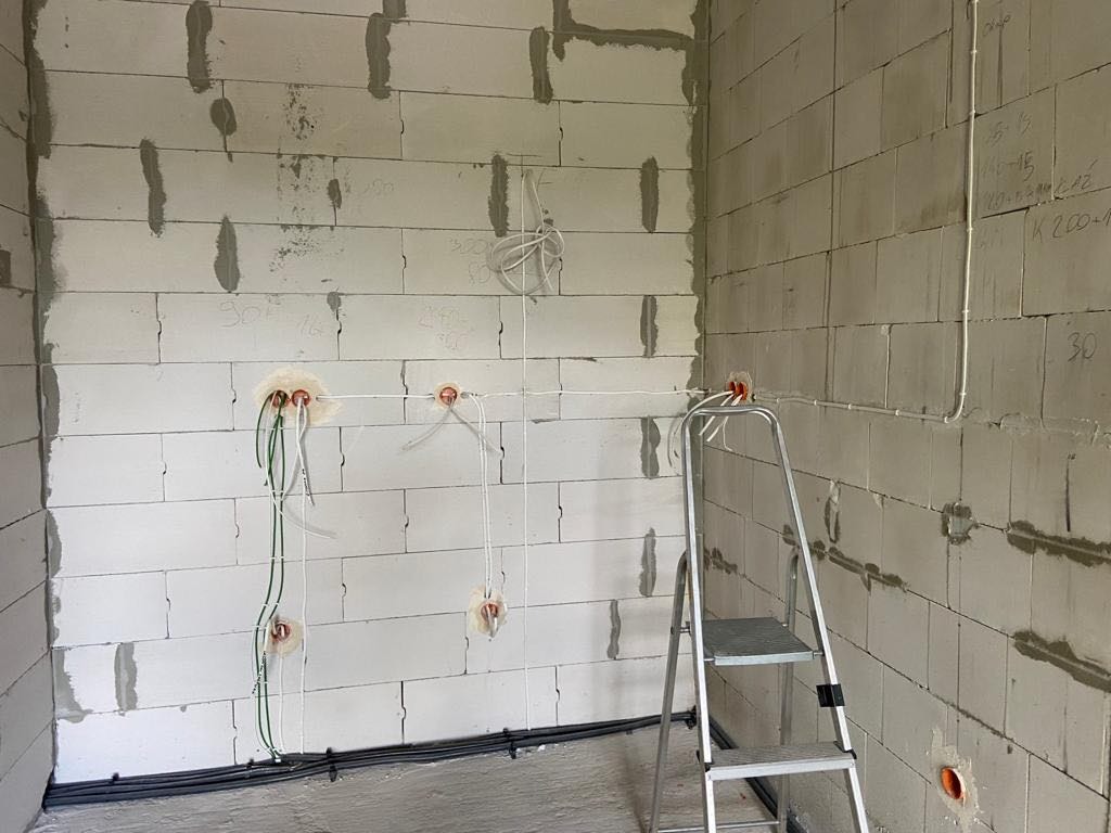 Wewnętrzna ściana z betonu komórkowego w trakcie prac elektrycznych: widoczne przewody w peszlach, puszki instalacyjne, drabina aluminiowa oparta o ścianę, oraz kable prowadzone wzdłuż podłogi.