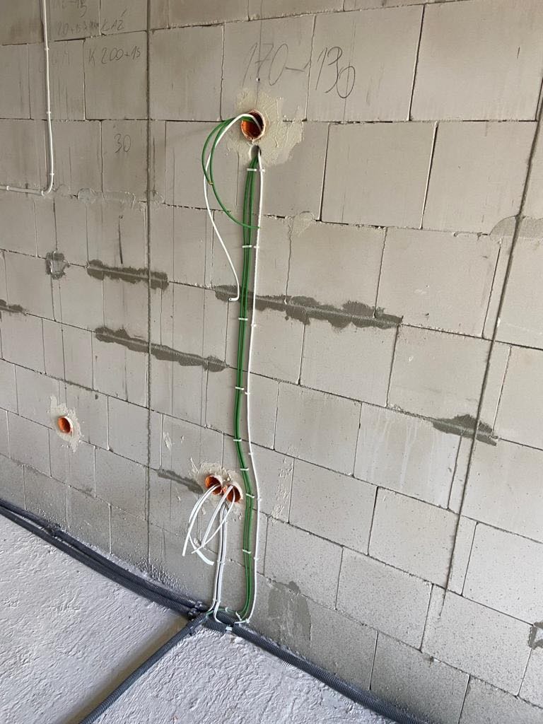 Ściana z betonu komórkowego z widocznymi przewodami elektrycznymi wychodzącymi z otworów w ścianie, zabezpieczonymi pomarańczowymi peszlami, oraz ułożonymi wzdłuż ściany w korytku podłogowym.