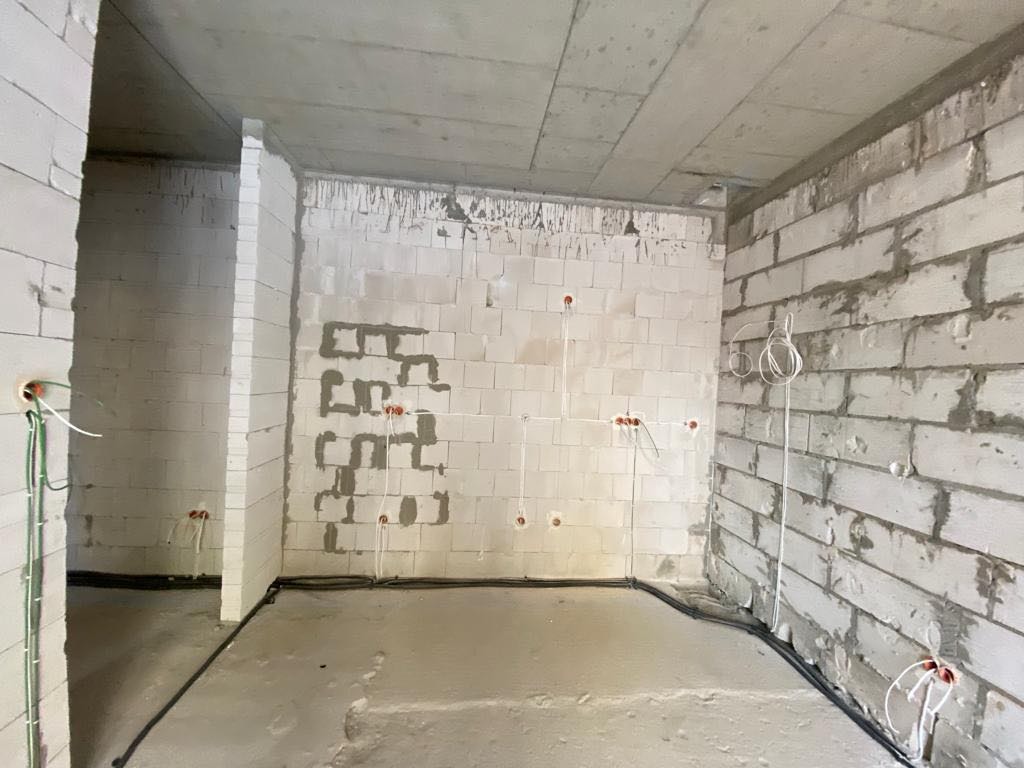 Surowy stan wnętrza z widocznymi przewodami elektrycznymi w ścianach z białego bloczka, z bruzdami pod instalację, oraz czarnymi kablami prowadzonymi po podłodze.