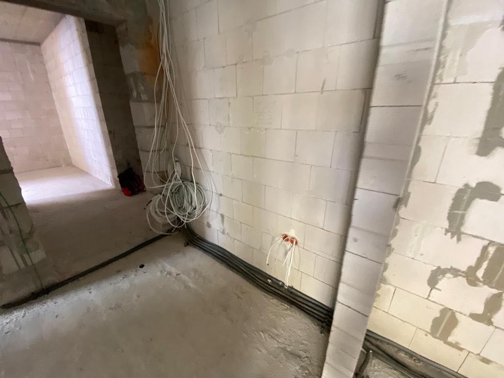 Surowy stan instalacji elektrycznej w nowym budynku: przewody zwisające ze ściany z bloczków, rury instalacyjne ułożone wzdłuż podłogi, widoczne puszki instalacyjne z wystającymi przewodami.