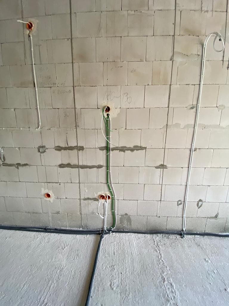 Wewnętrzne okablowanie elektryczne w surowym stanie, przewody białe i zielone wychodzące z otworów w ścianie z betonu komórkowego, widoczne czarne rury przy podłodze.