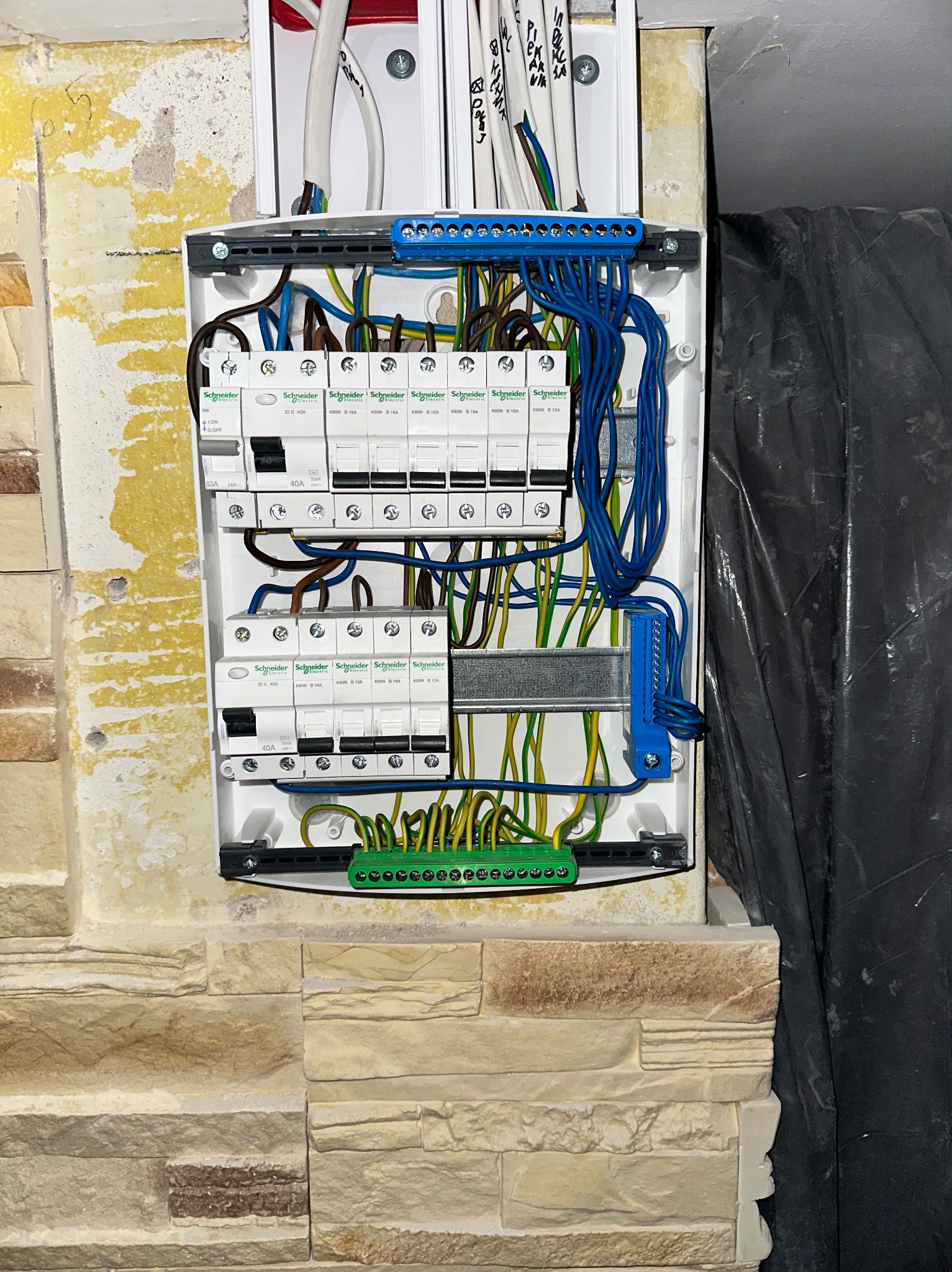 Rozdzielnica elektryczna Schneider z podłączonymi przewodami w kolorach: niebieskim, brązowym i żółto-zielonym. Widoczne zabezpieczenia. Tło stanowi ściana z dekoracyjnej cegły.