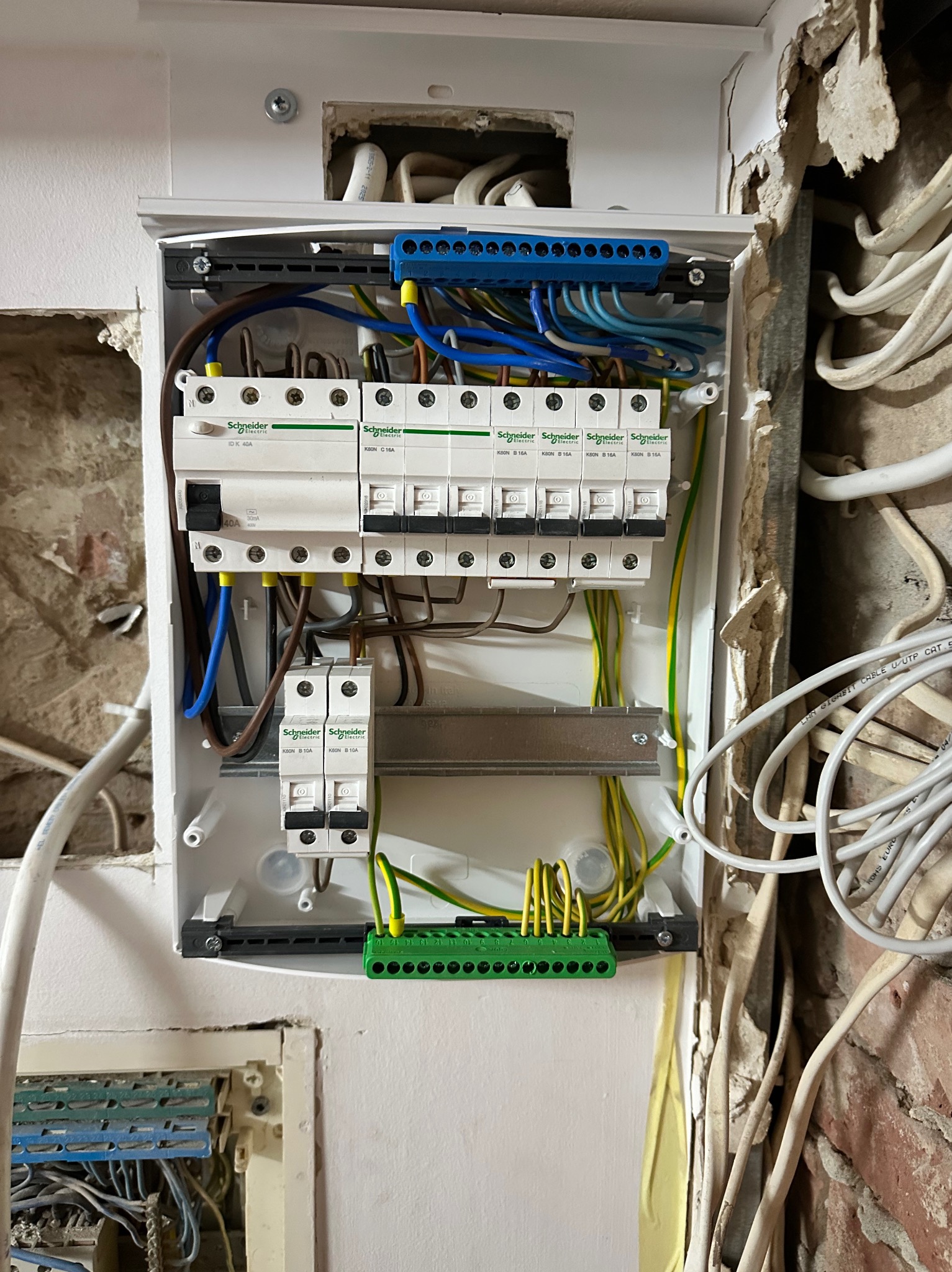 Rozdzielnica elektryczna Schneider z podłączonymi przewodami w trakcie montażu, widoczne zabezpieczenia i listwy zaciskowe. Ściana z uszkodzonym tynkiem.