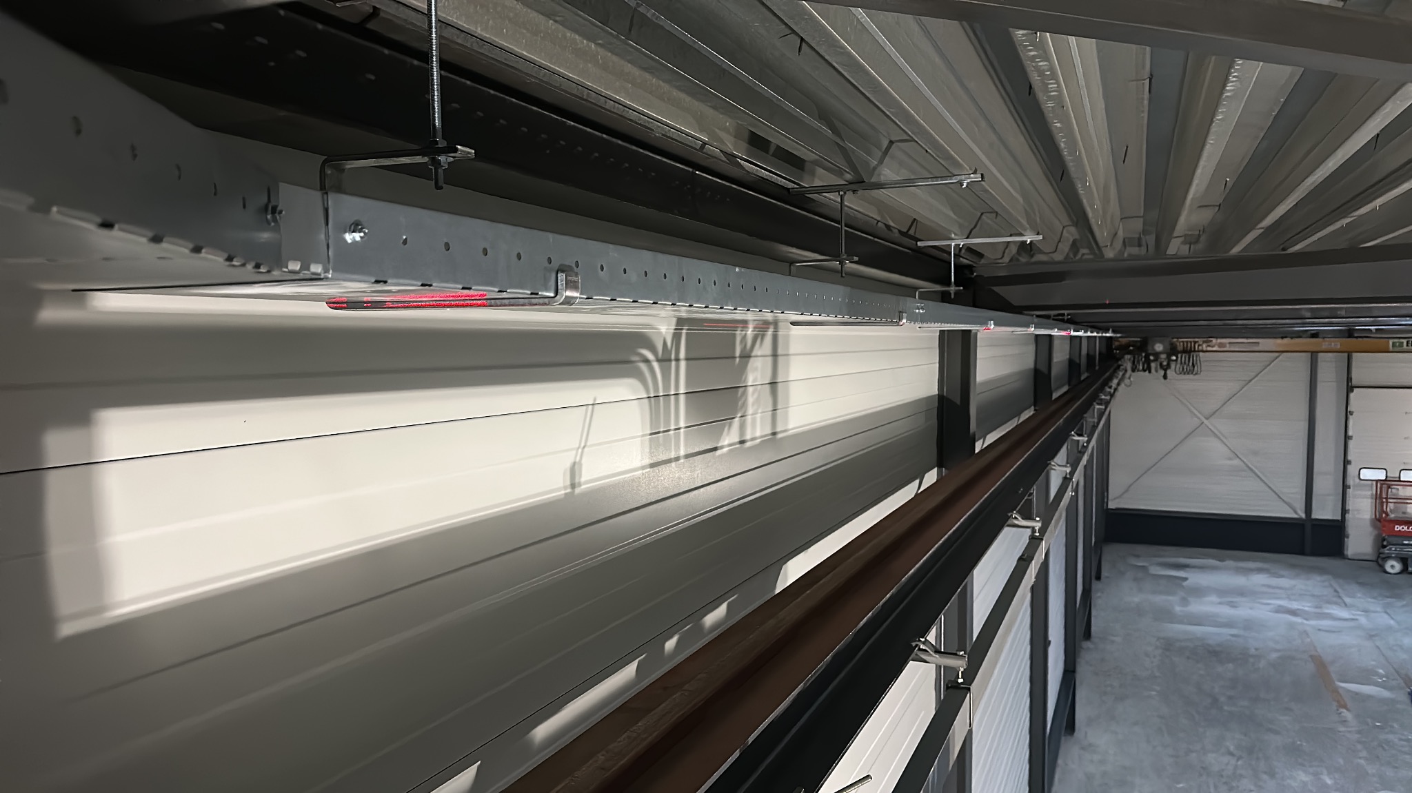 Szara, metalowa konstrukcja wsporcza pod sufitem hali, prawdopodobnie trasa kablowa. Widoczne elementy mocujące i fragment instalacji. Oświetlenie hali.