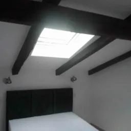 Sypialnia z białymi ścianami, ciemnymi belkami stropowymi, oknem dachowym i łóżkiem z ciemnozielonym zagłówkiem.