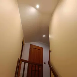 Wnętrze klatki schodowej z drewnianą barierką, świeżo pomalowanymi ścianami w odcieniach beżu i widocznymi lampami sufitowymi.