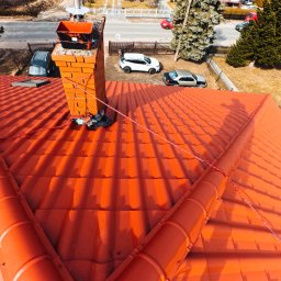 Art.Male - Widok z góry na dach pokryty czerwoną dachówką, z widocznym kominem z cegły, na którym stoi wiadro i leżą narzędzia, w tle ulica z zaparkowanymi samochodami i drzewa.