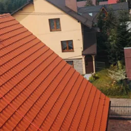 Nowy, pomarańczowy dach z dachówki ceramicznej na tle domu z beżową elewacją i górskiego krajobrazu, widoczna lina zabezpieczająca.