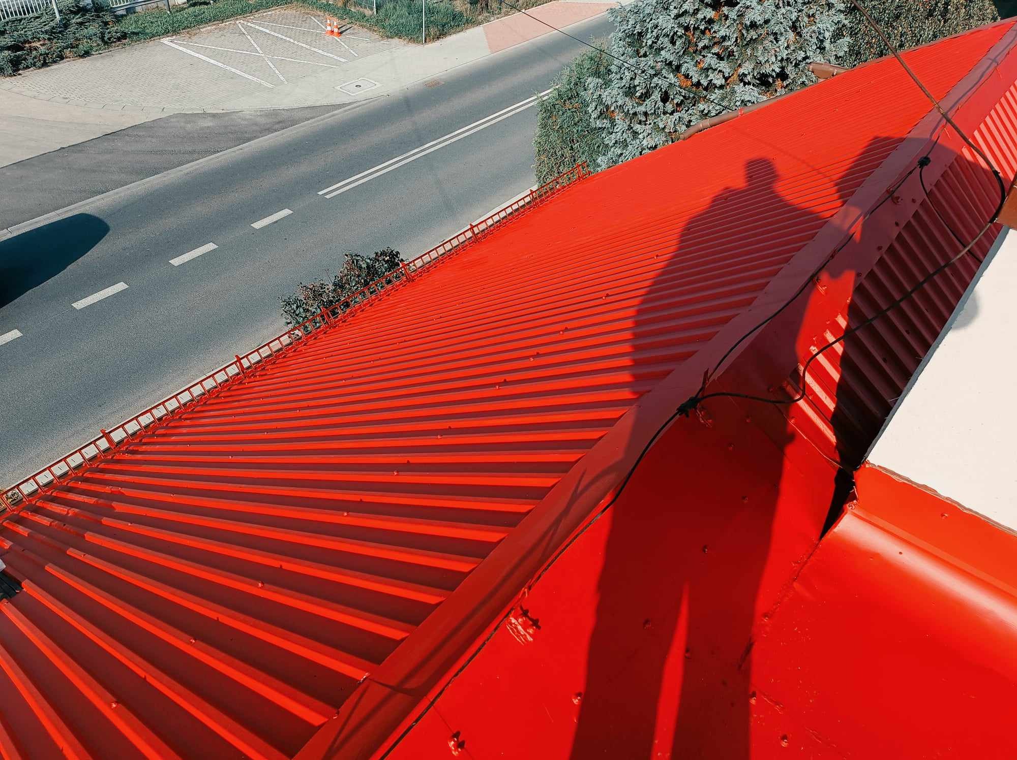 Czerwony, blaszany dach po renowacji, widok z góry na ulicę i cień osoby pracującej na dachu.