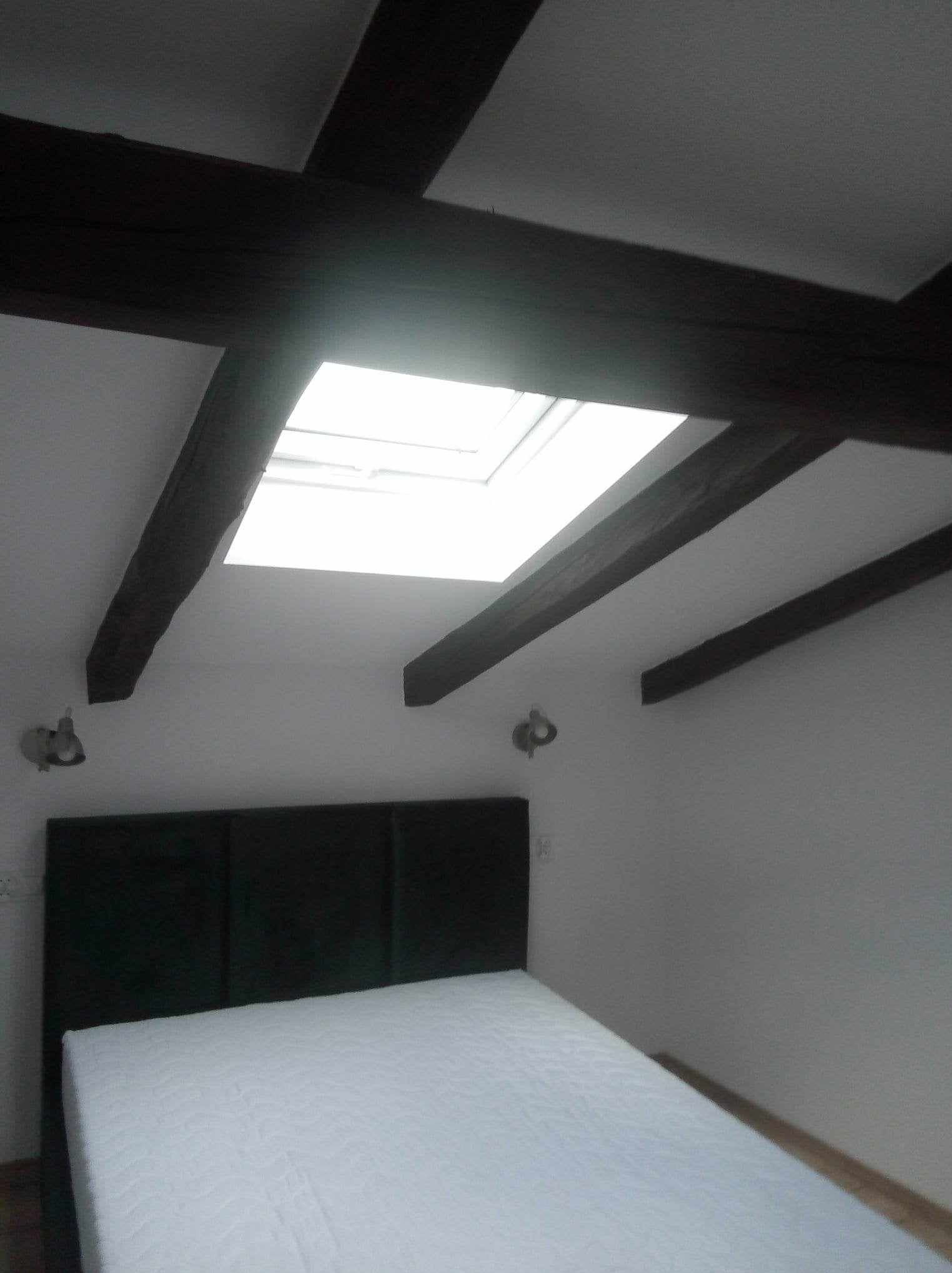 Sypialnia z białymi ścianami, ciemnymi belkami stropowymi, oknem dachowym i łóżkiem z ciemnozielonym zagłówkiem.