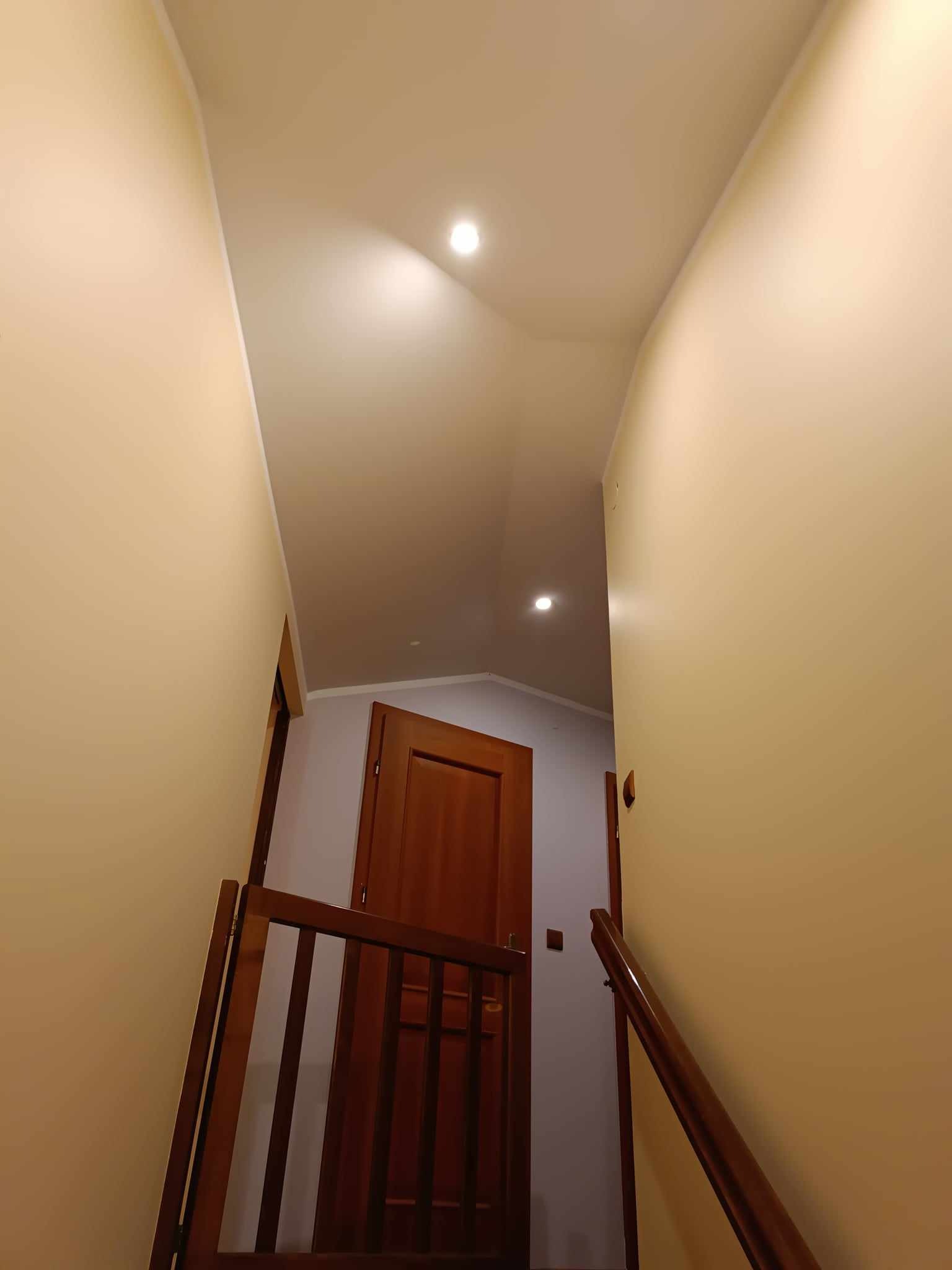 Wnętrze klatki schodowej z drewnianą barierką, świeżo pomalowanymi ścianami w odcieniach beżu i widocznymi lampami sufitowymi.