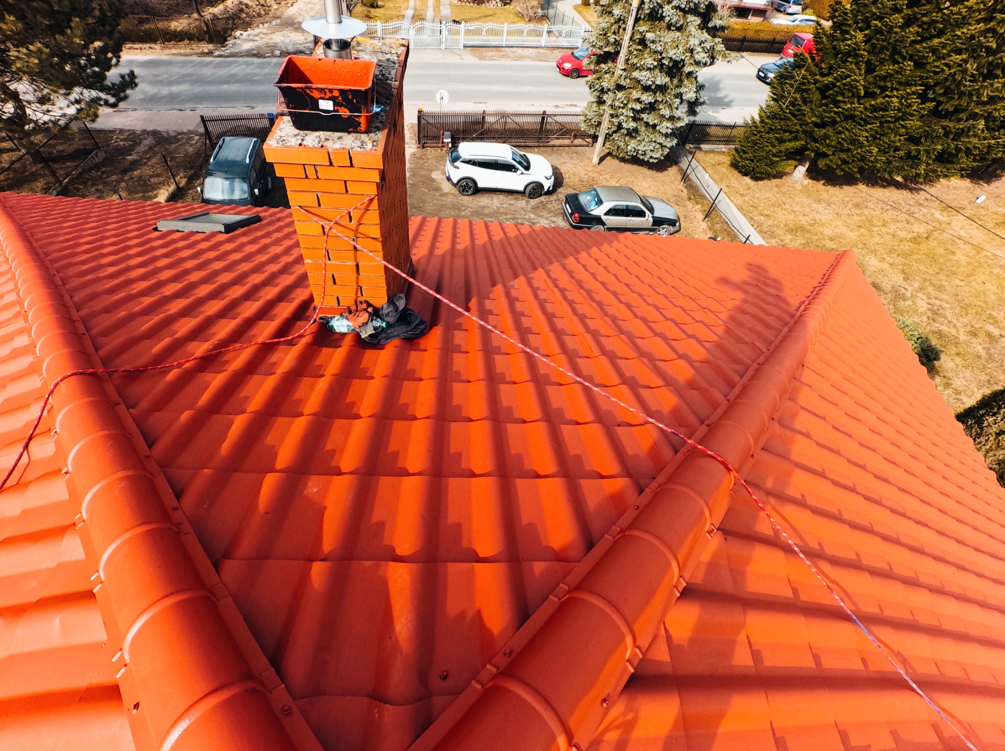 Widok z góry na dach pokryty czerwoną dachówką, z widocznym kominem z cegły, na którym stoi wiadro i leżą narzędzia, w tle ulica z zaparkowanymi samochodami i drzewa.