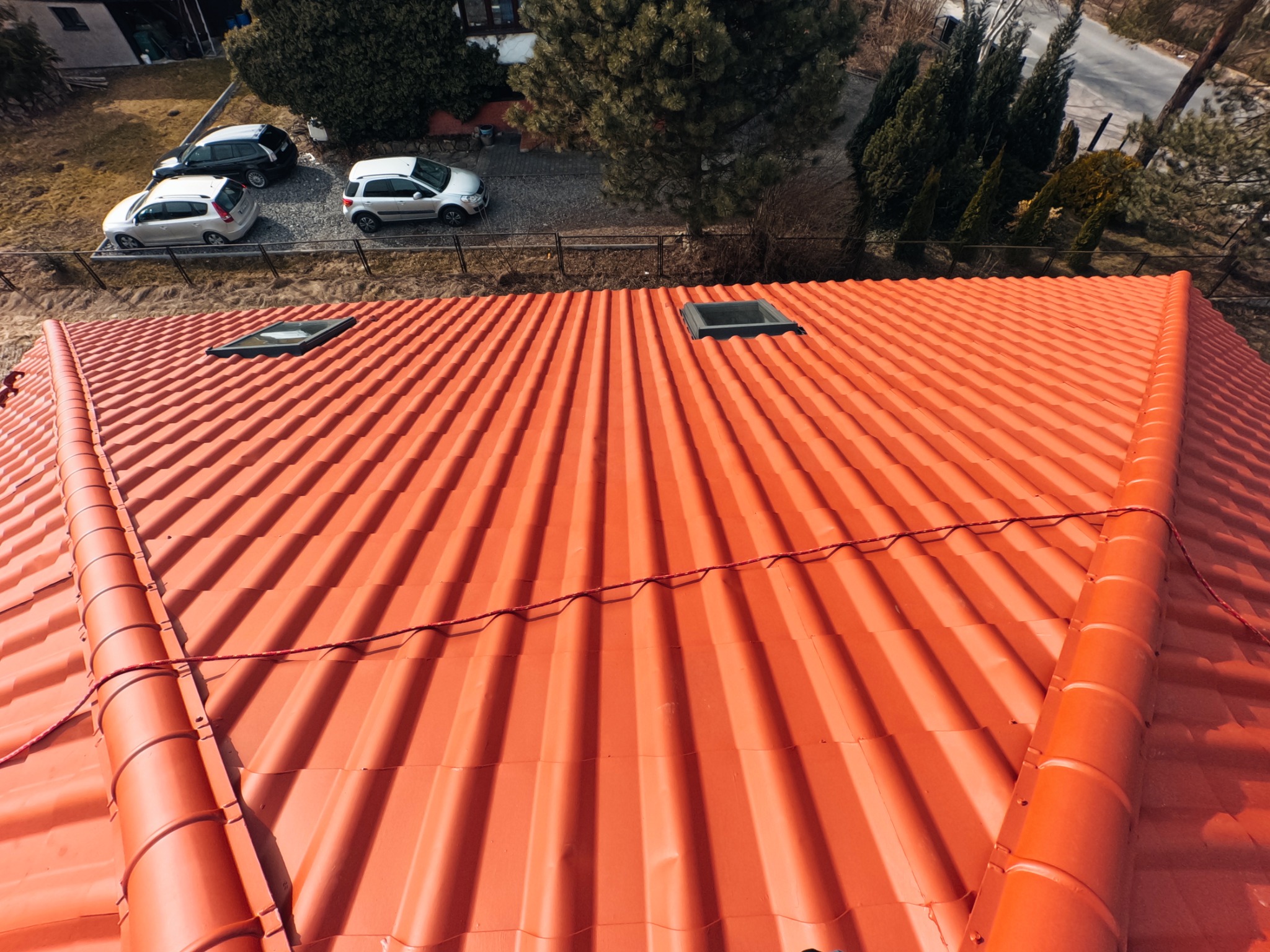 Widok z góry na nowy, pomarańczowy dach z blachodachówki z dwoma oknami dachowymi i widocznymi samochodami na podjeździe.