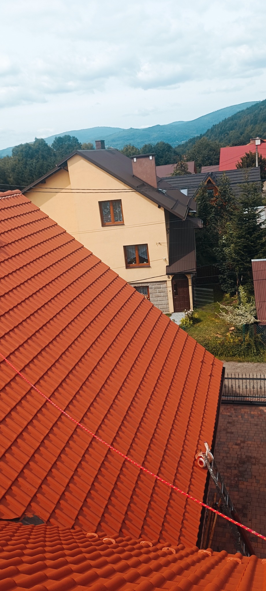 Nowy, pomarańczowy dach z dachówki ceramicznej na tle domu z beżową elewacją i górskiego krajobrazu, widoczna lina zabezpieczająca.