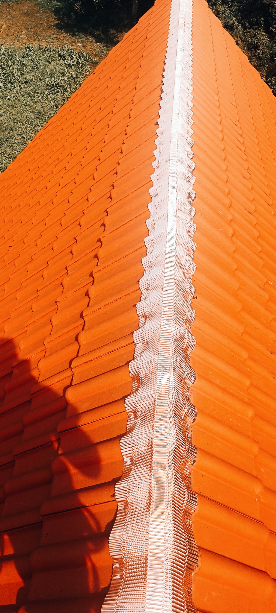 Pomarańczowy dach z dachówki ceramicznej z wykończeniem kalenicy w kolorze srebrnym, widziany z góry pod kątem, z fragmentem zieleni w tle.