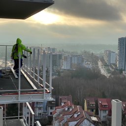 GLOBALSKILL SPÓŁKA Z OGRANICZONĄ ODPOWIEDZIALNOŚCIĄ - Praca na balkonie: monter w odblaskowej kurtce instaluje metalową balustradę na tle miejskiego krajobrazu w pochmurny dzień. Widoczne narzędzia i elementy konstrukcyjne.