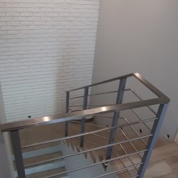 GLOBALSKILL SPÓŁKA Z OGRANICZONĄ ODPOWIEDZIALNOŚCIĄ - Metalowa balustrada schodowa z poziomymi prętami, widok z góry, biała ściana z cegły w tle, drewnopodobne płytki na podłodze, nowoczesny design.